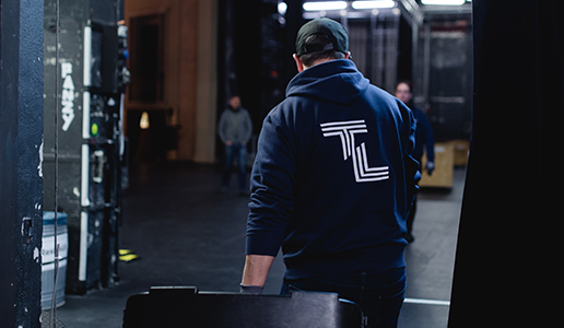 Blick hinter die Bühne: Ein Techniker mit TL-Hoodie schiebt einen Transportkoffer durch den Bühnenbereich während des Aufbaus.