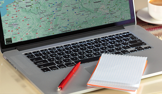 Laptop mit geöffneter Landkarte, daneben ein roter Stift, ein kariertes Notizbuch und ein Kaffee – Symbol für die Arbeit im Büro bei Theater Liberi.